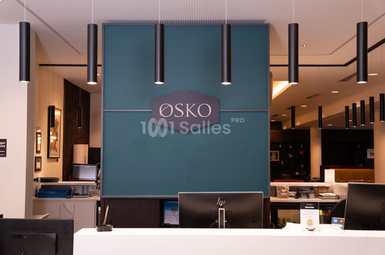Comptoir d'accueil moderne avec un panneau ’ØSKO’ sur un mur bleu et des luminaires suspendus noirs.