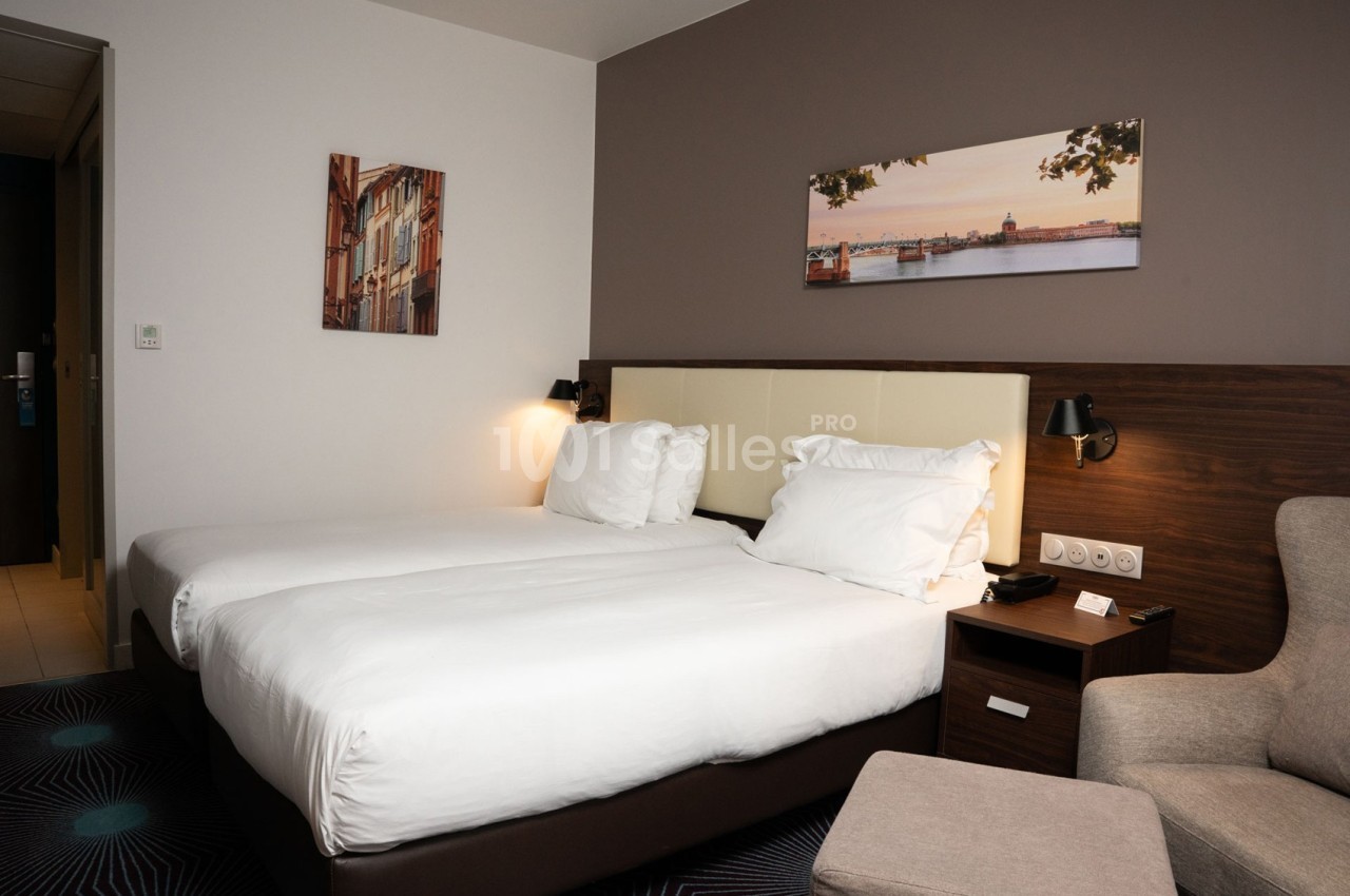 Chambre d'hôtel avec deux lits simples, décorée de tableaux muraux et équipée d'un fauteuil et d'une table de chevet.