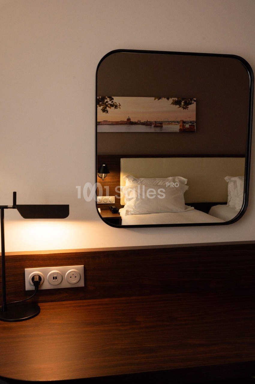 Bureau en bois avec lampe, miroir reflétant un lit et un tableau mural dans une chambre sobrement éclairée.