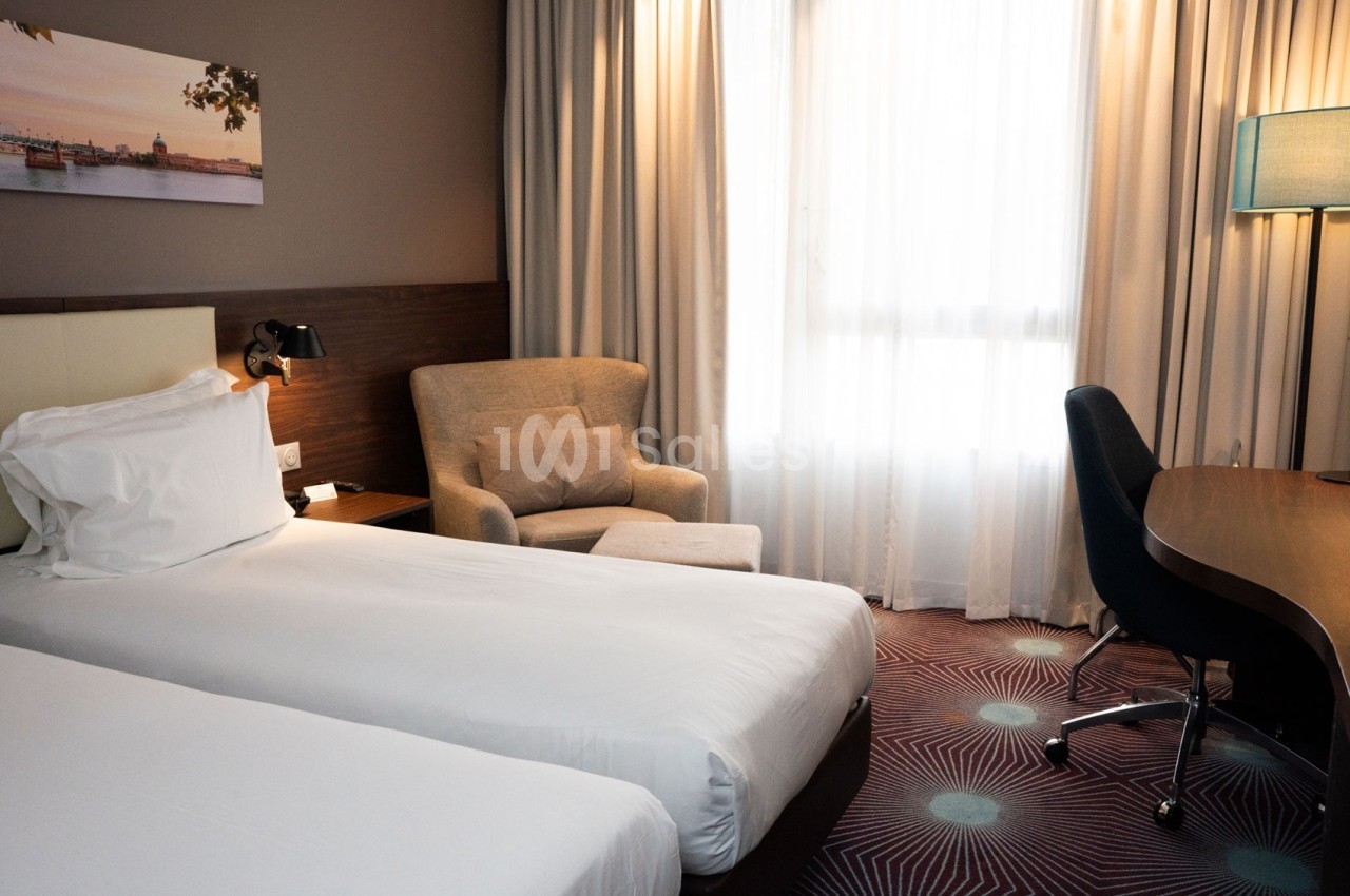 Chambre d'hôtel avec deux lits simples, un fauteuil, un bureau, une lampe et une grande fenêtre avec rideaux.