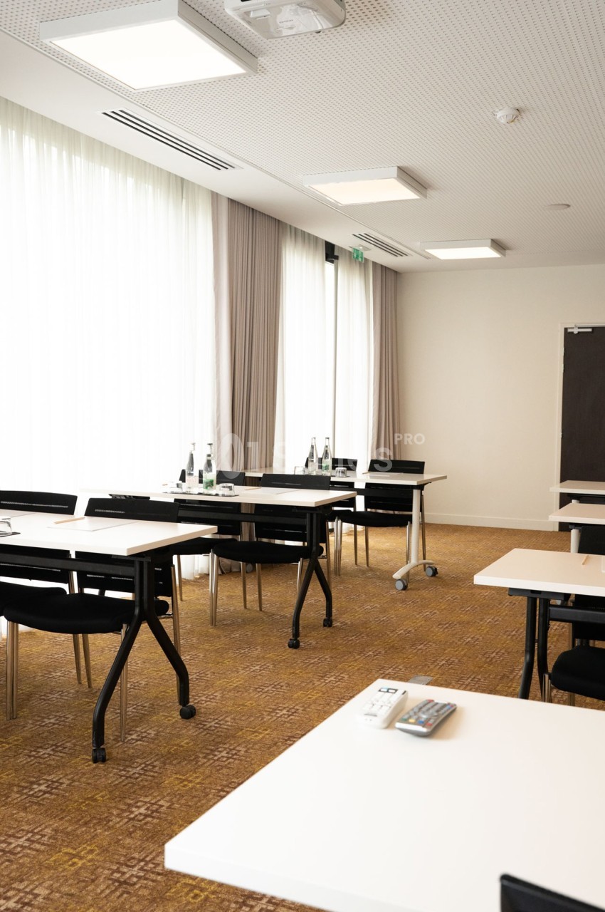 Salle de réunion lumineuse avec tables blanches, chaises noires, bouteilles d'eau et rideaux beige clair.
