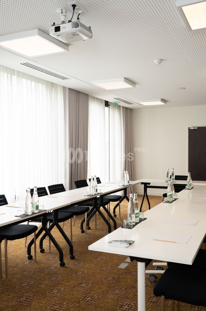 Salle de réunion lumineuse avec tables blanches disposées en U, chaises noires et bouteilles d'eau sur les tables.