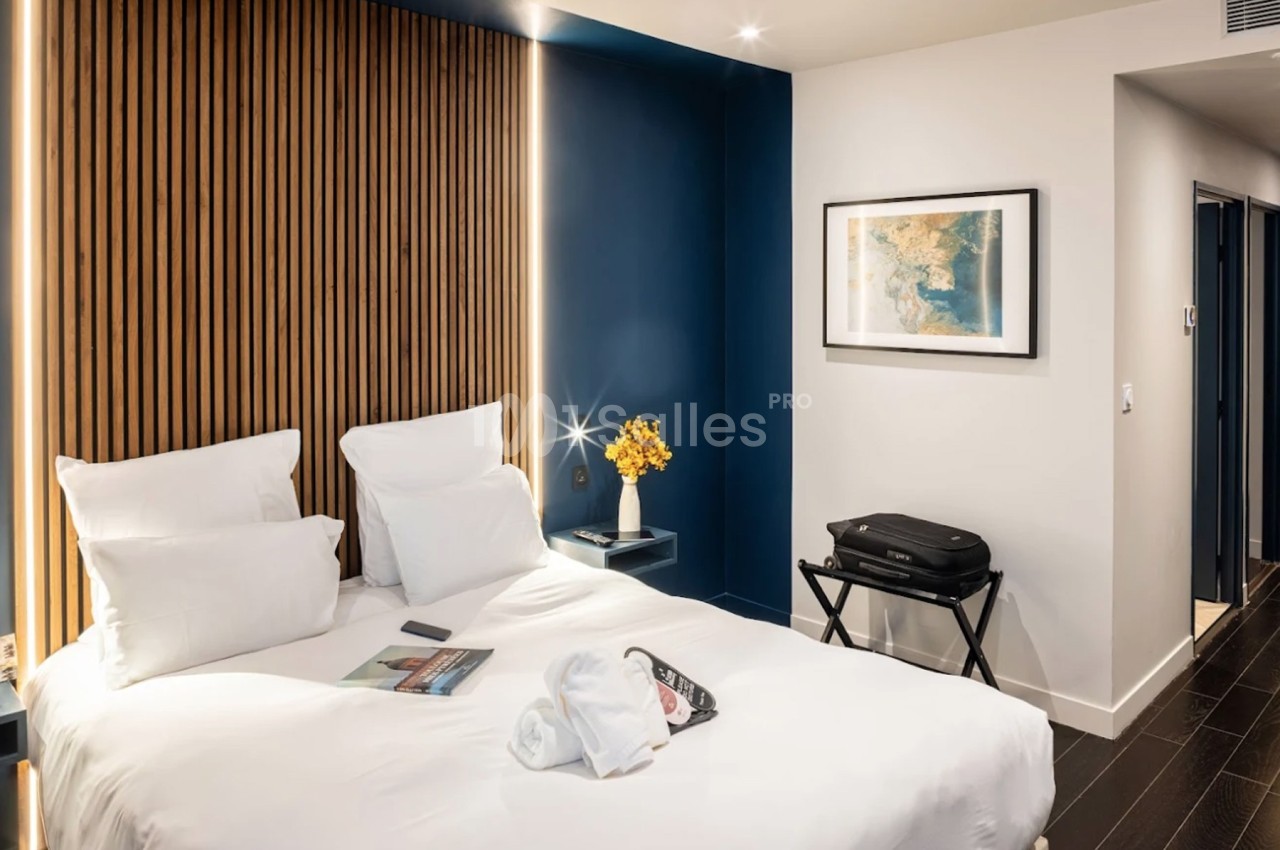 Chambre moderne avec lit double, mur bleu, décoration minimaliste, valise sur support et lumière douce.
