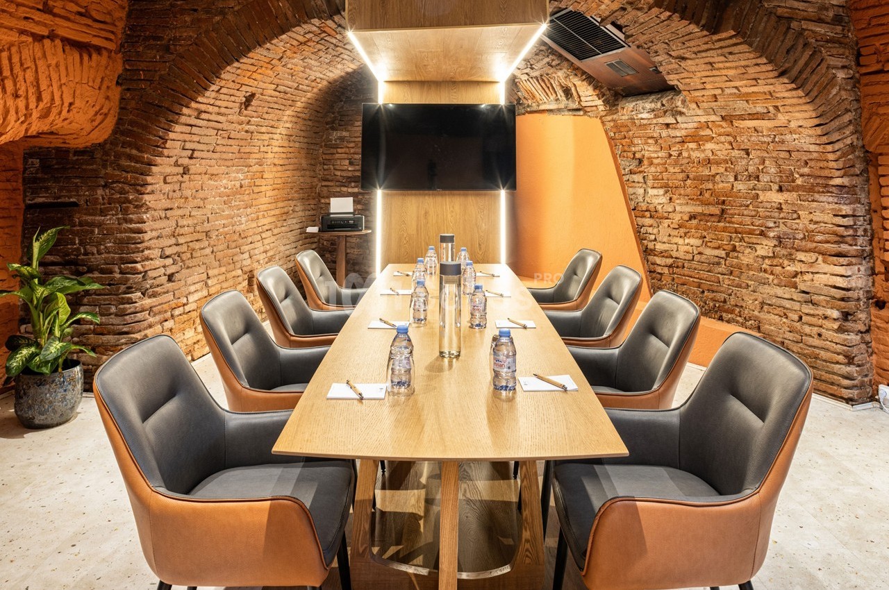 Salle de réunion voûtée en briques avec table en bois, chaises modernes, écran mural et bouteilles d'eau alignées.