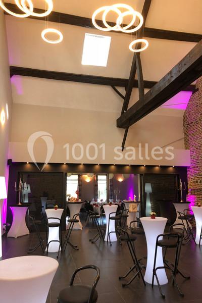 Salle de réception avec tables hautes, chaises noires, éclairage moderne et poutres apparentes sous un plafond haut.