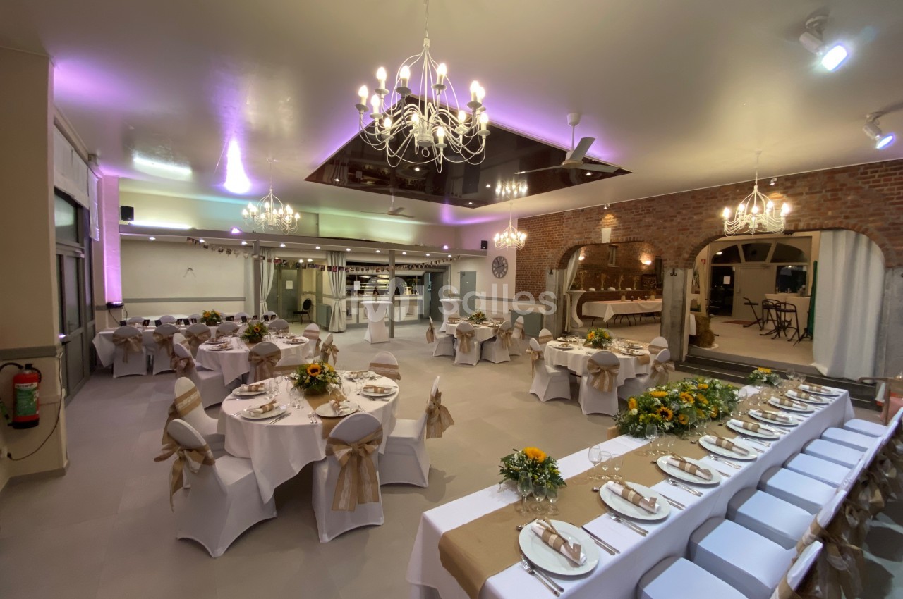 Salle de réception décorée avec des tables rondes et rectangulaires, nappes blanches, fleurs jaunes et éclairage chaleureux.