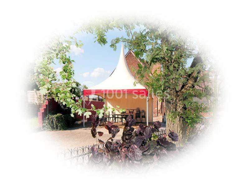 Tente blanche avec toit pointu et bordure rouge installée dans un jardin arboré, entourée de tables et chaises.