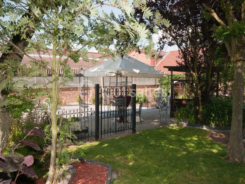 Jardin avec pelouse, arbres, pergola et parasol blanc, entouré de murs en briques et clôtures métalliques.