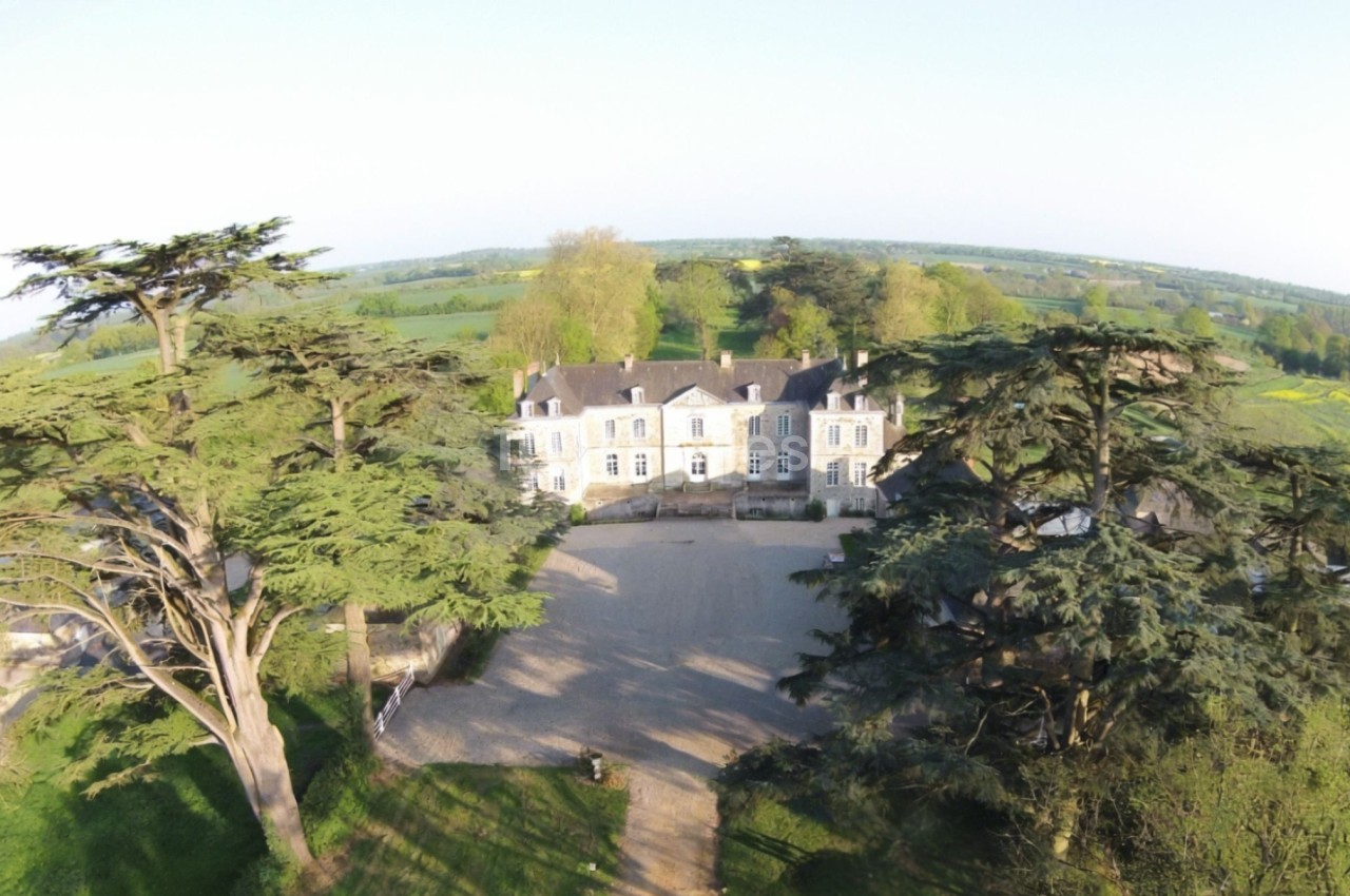 Vue aérienne d'un manoir entouré d'arbres et de champs verdoyants sous un ciel dégagé.