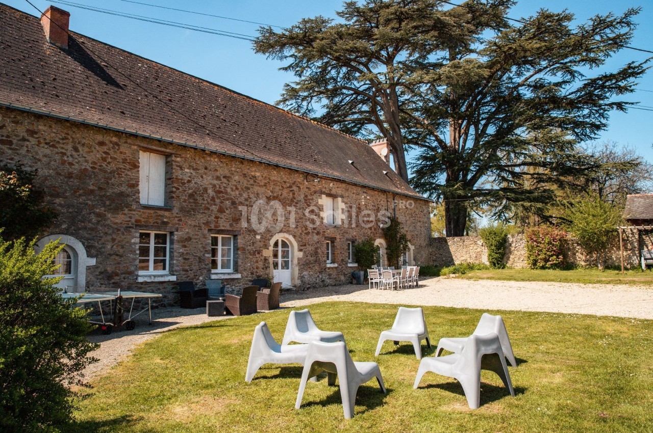 Cour d'une maison en pierre avec des chaises en plastique disposées sur une pelouse, entourée d'arbres et de végétation.