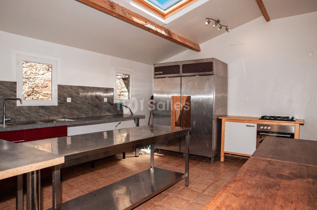Cuisine professionnelle avec plan de travail en inox, réfrigérateur industriel et éclairage naturel par une fenêtre de toit.