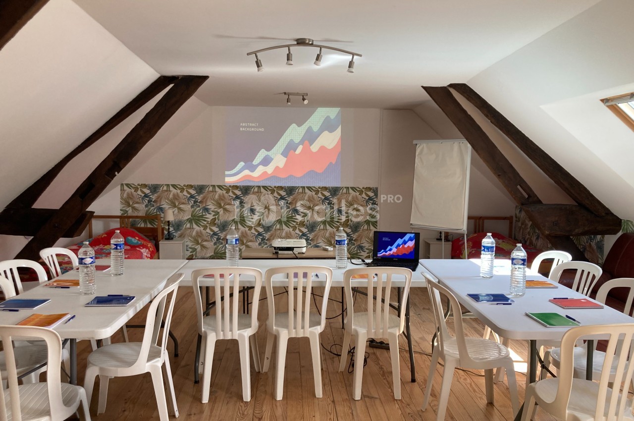 Salle de réunion lumineuse avec tables, chaises blanches, projecteur, écran, bloc-notes et bouteilles d'eau.