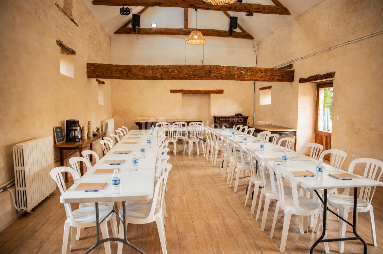 Salle de réunion lumineuse avec tables alignées, chaises blanches en plastique et poutres apparentes au plafond.