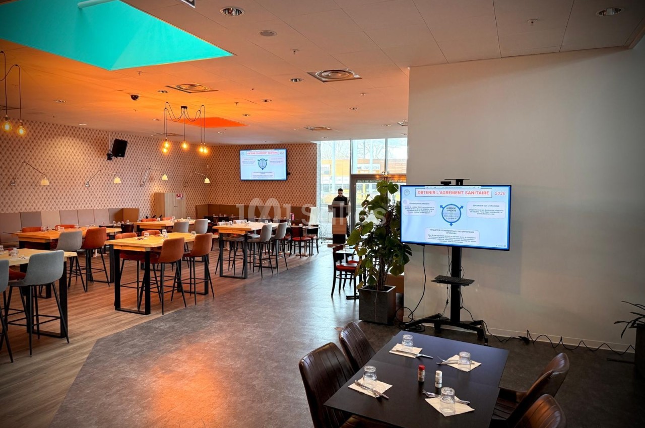 Salle de restaurant lumineuse avec tables dressées, écran de présentation et éclairage moderne.