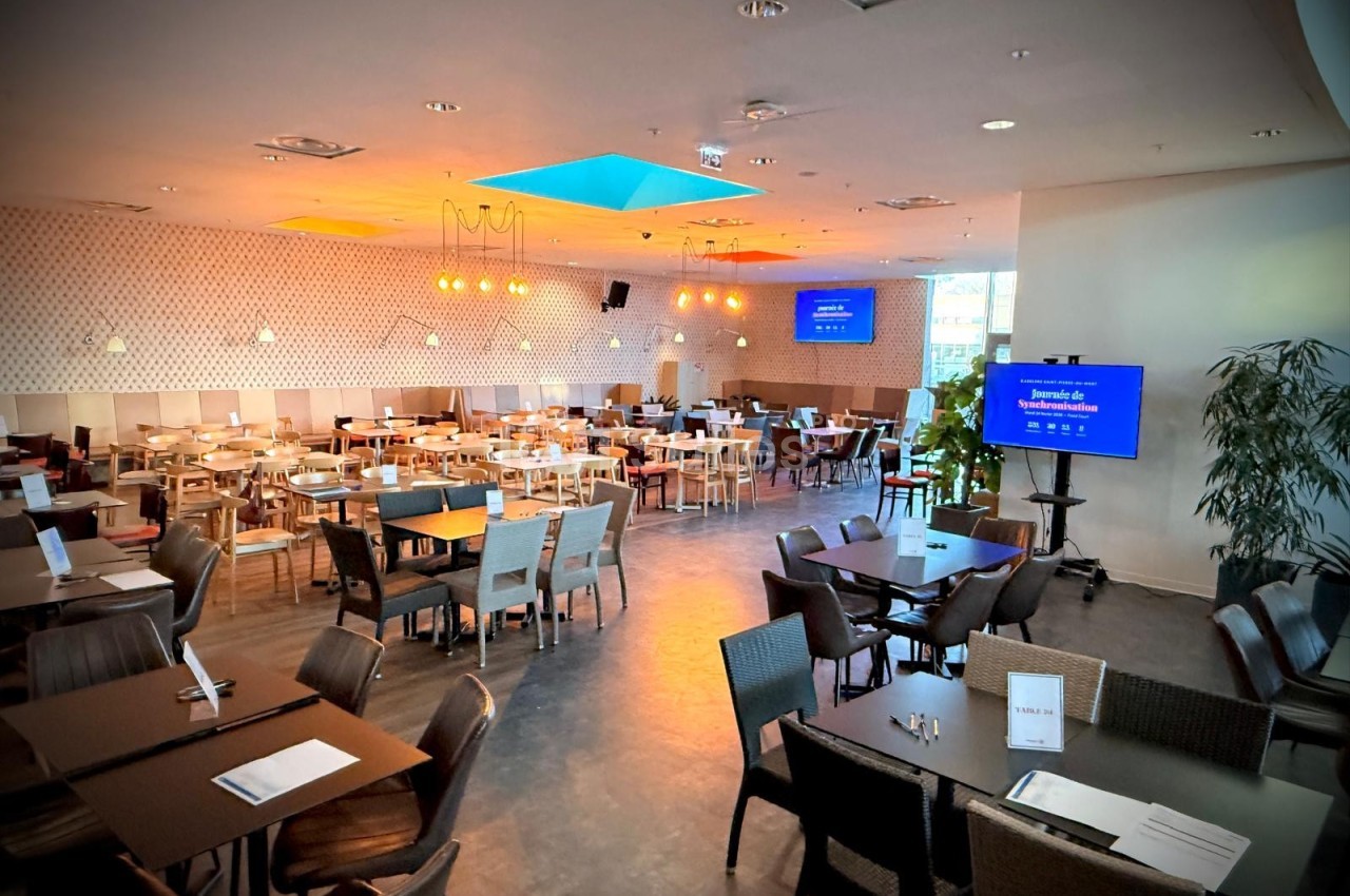 Salle de restaurant spacieuse avec tables et chaises disposées, éclairage chaleureux et écran de présentation allumé.