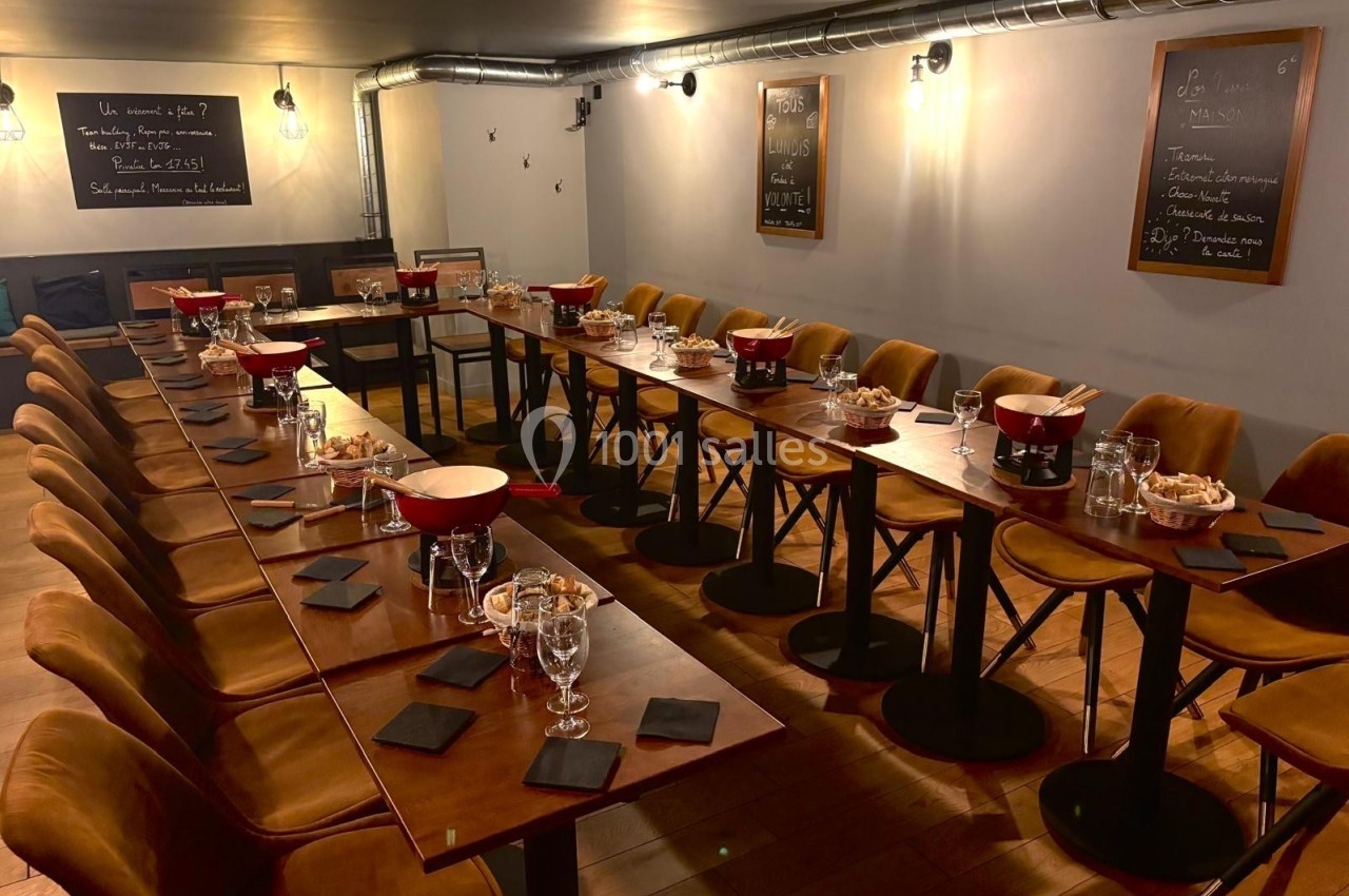 Salle de restaurant aménagée avec de longues tables en bois, chaises rembourrées et vaisselle prête pour un repas.