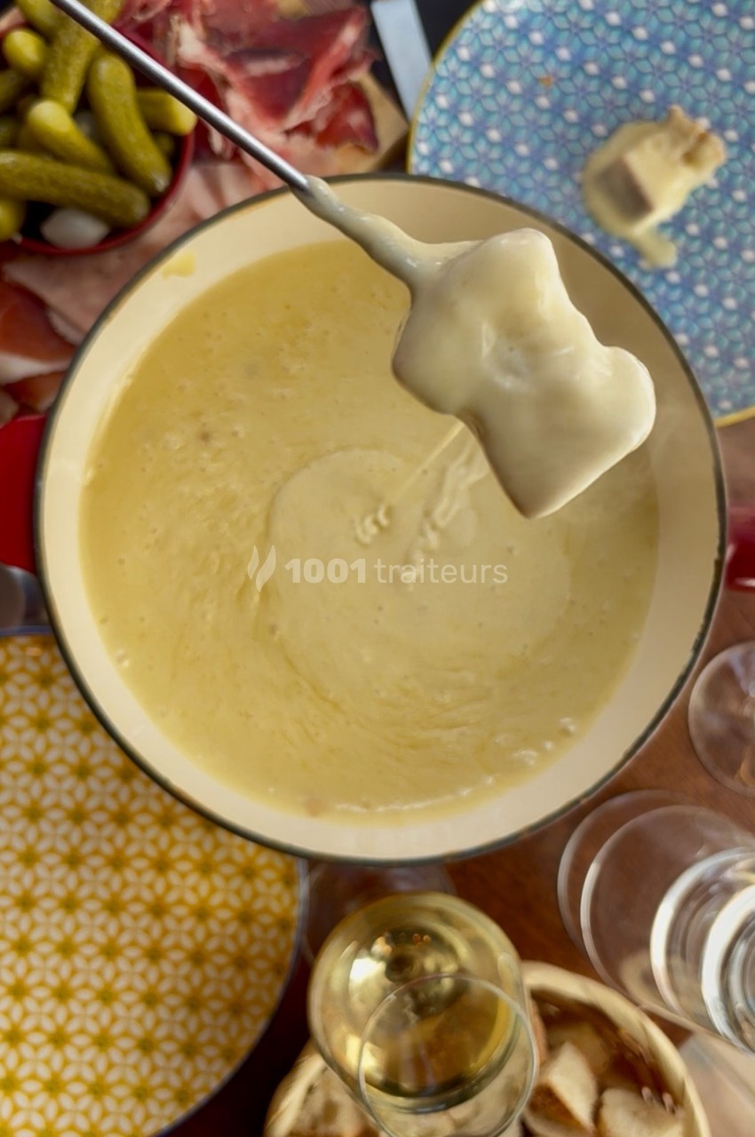 Fromage fondu dans une casserole avec un morceau de pain trempé, entouré de charcuterie, cornichons et assiettes colorées.