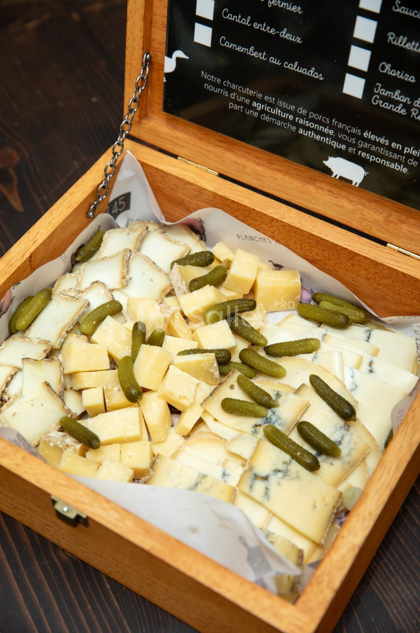 Plateau en bois contenant des cubes de fromage variés et des cornichons, présenté sur un fond sombre.