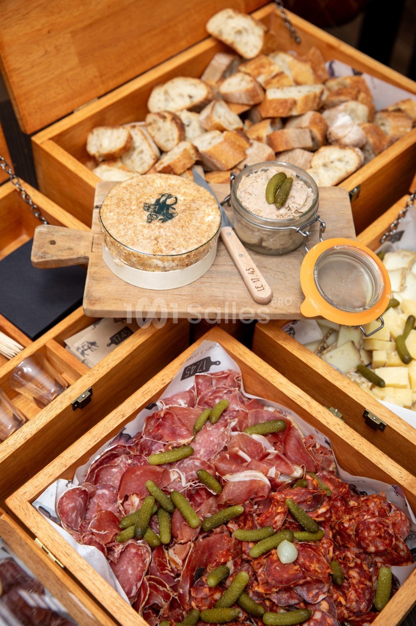 Plateau de charcuterie avec cornichons, fromage, pain tranché et rillettes présentés dans des boîtes en bois.