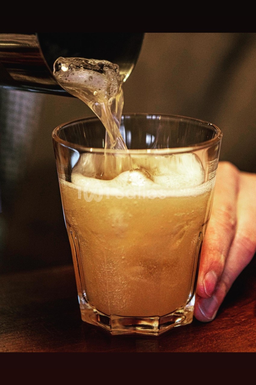 Un liquide mousseux est versé dans un verre transparent tenu par une main sur une table en bois.
