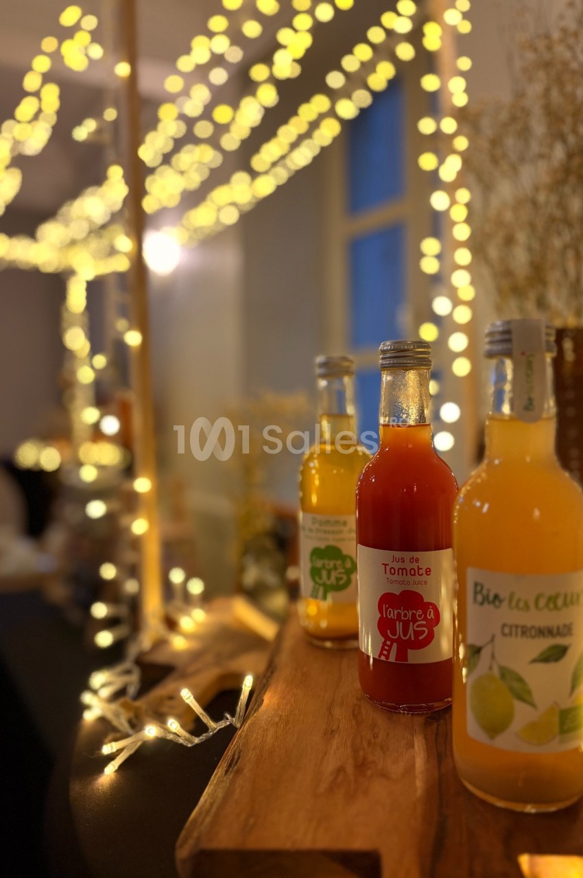Bouteilles de jus artisanaux disposées sur un support en bois, éclairées par des guirlandes lumineuses dans un décor…