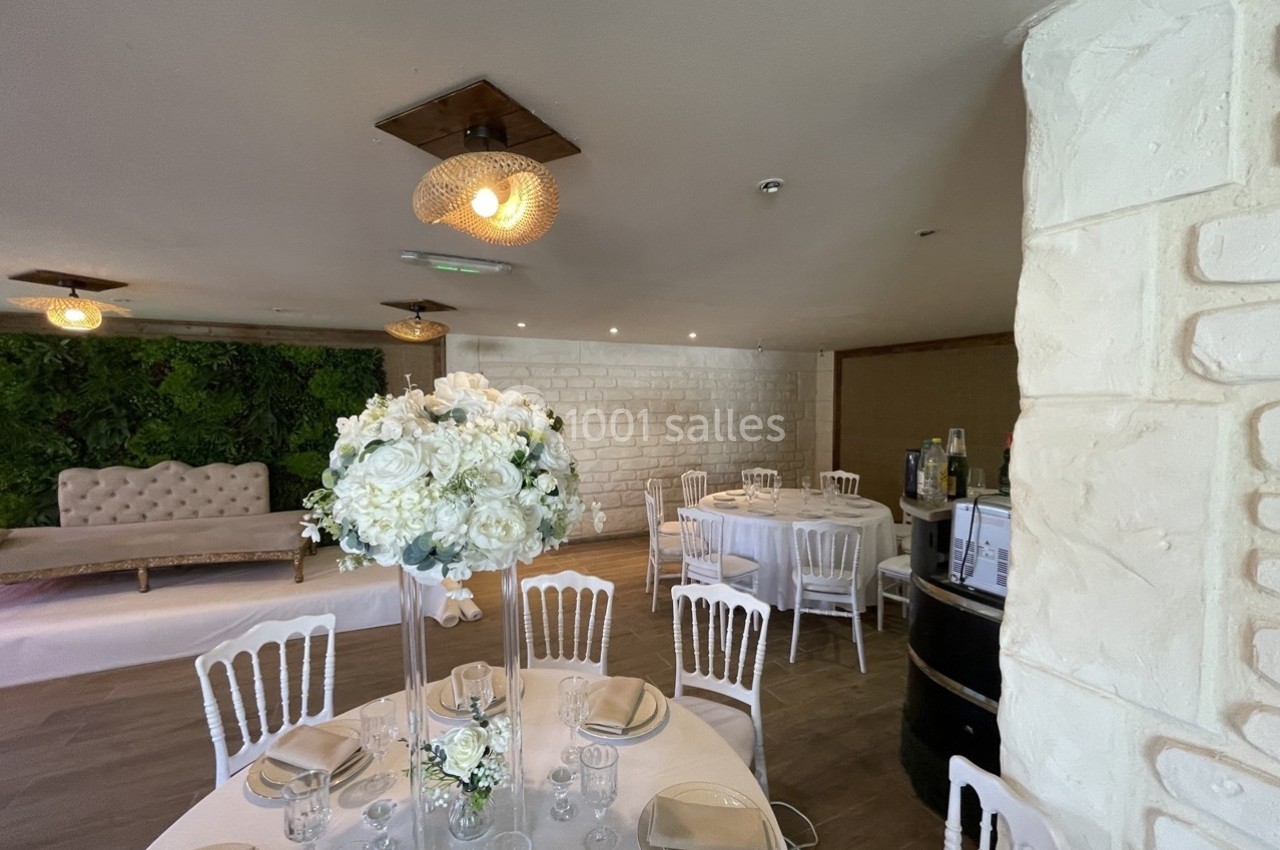 Salle de réception décorée avec des tables rondes, des chaises blanches et un bouquet de fleurs blanches au centre.