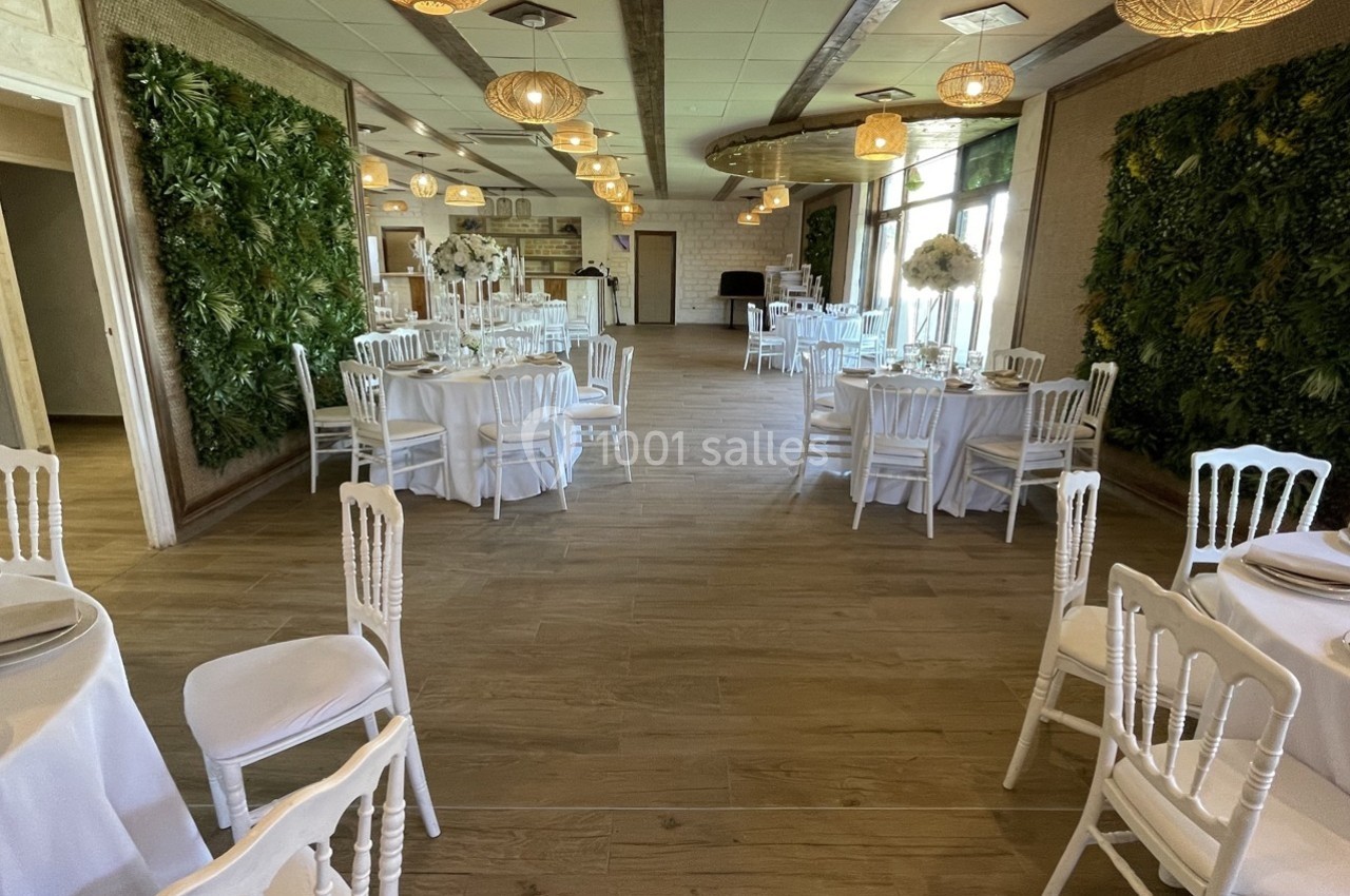 Salle de réception lumineuse avec tables rondes dressées, chaises blanches et murs végétalisés.