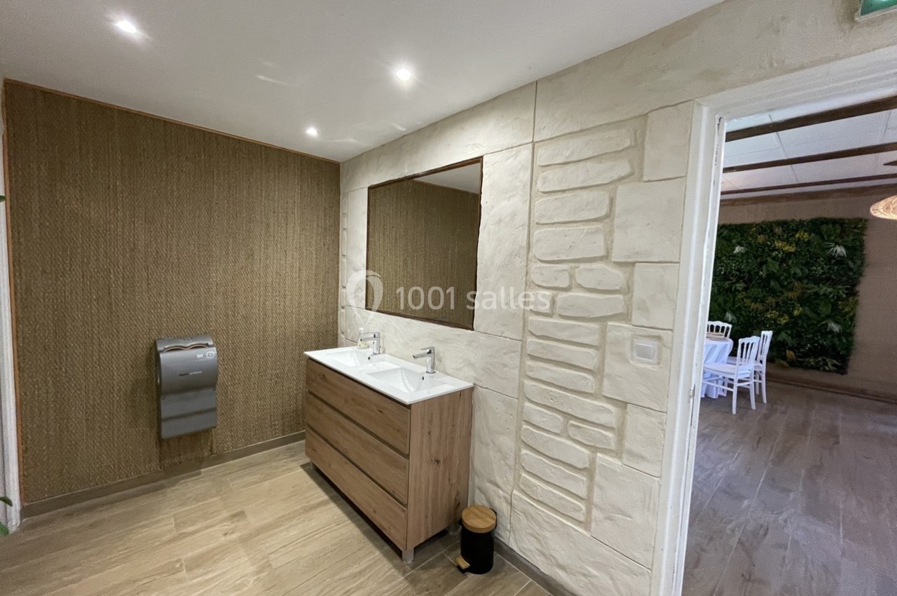 Salle de bain avec double vasque, miroir, mur en pierre et accès ouvert sur une pièce avec des chaises blanches.