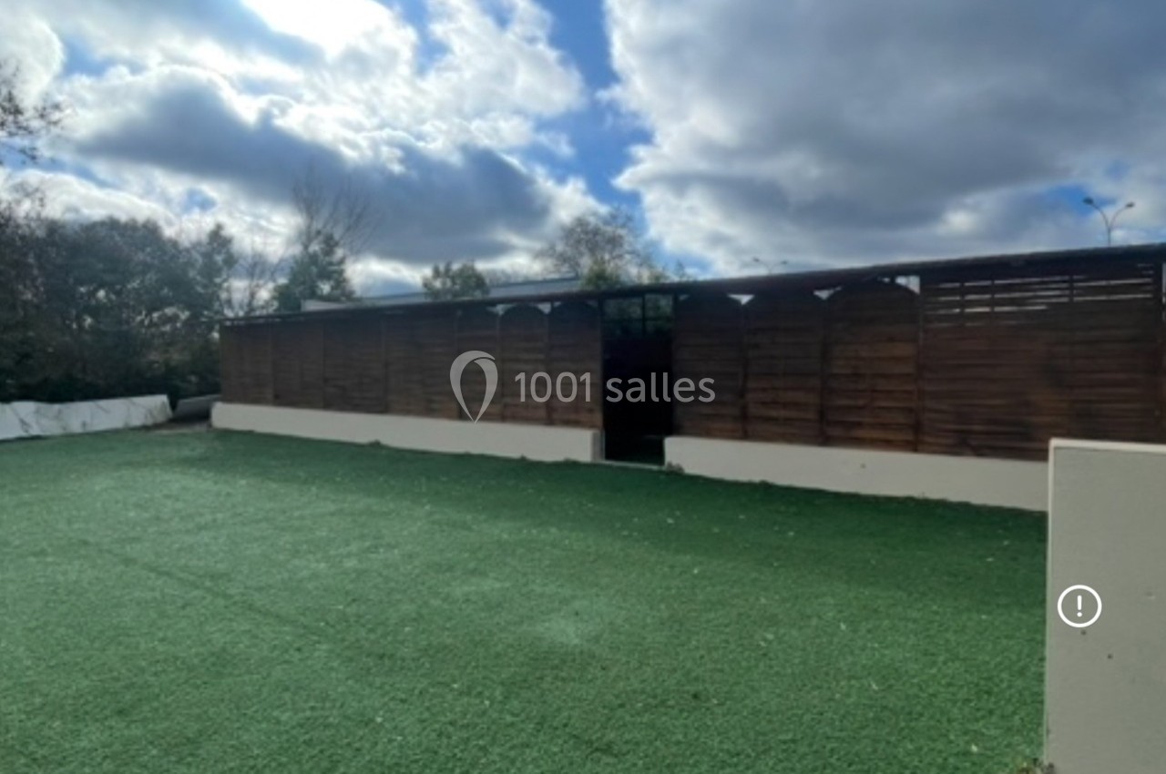Un bâtiment en bois avec une clôture, entouré d'une pelouse artificielle sous un ciel nuageux.