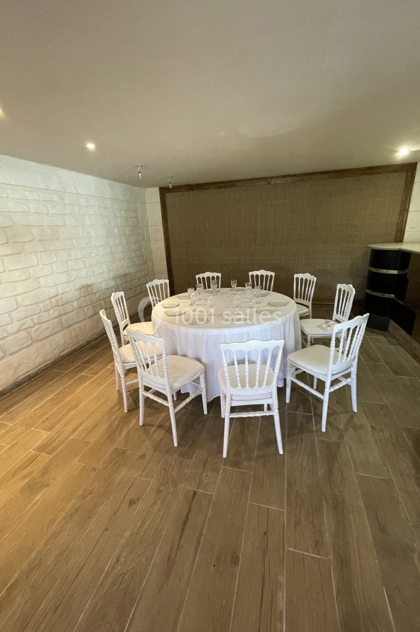 Table ronde dressée avec nappe blanche et chaises blanches dans une salle au sol en bois et murs clairs.