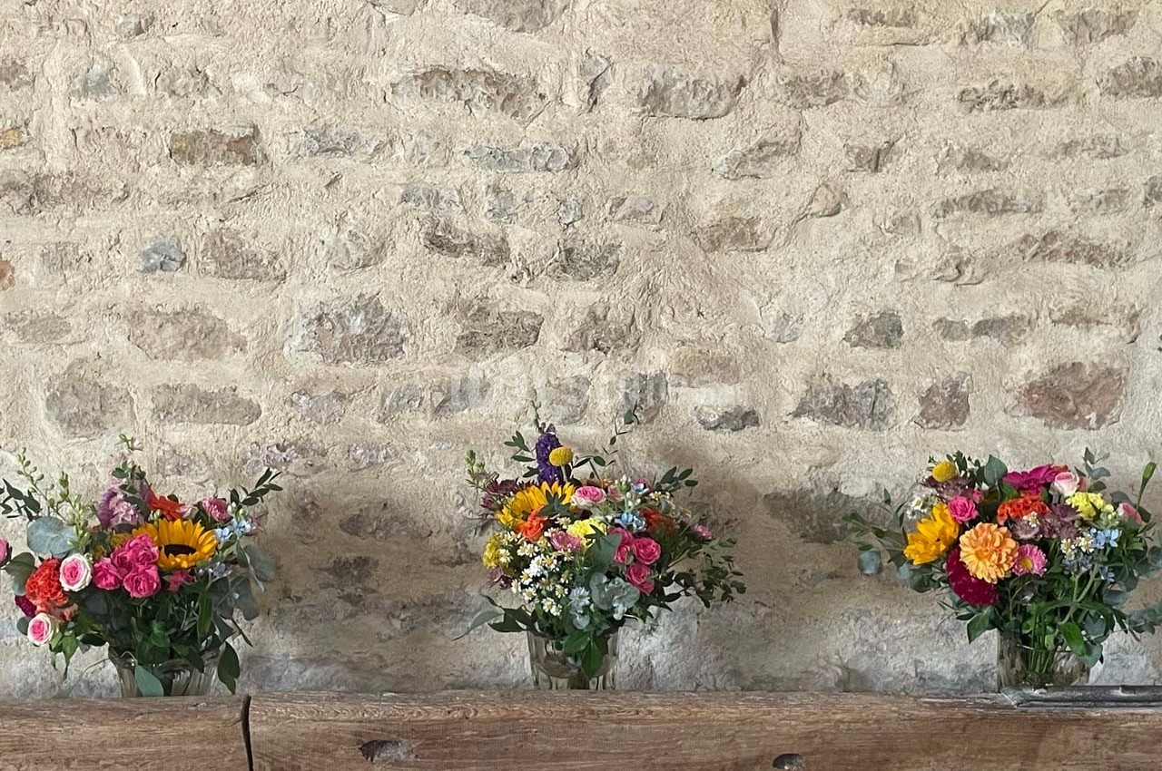 Trois bouquets de fleurs colorées posés sur une table en bois devant un mur en pierre brute.