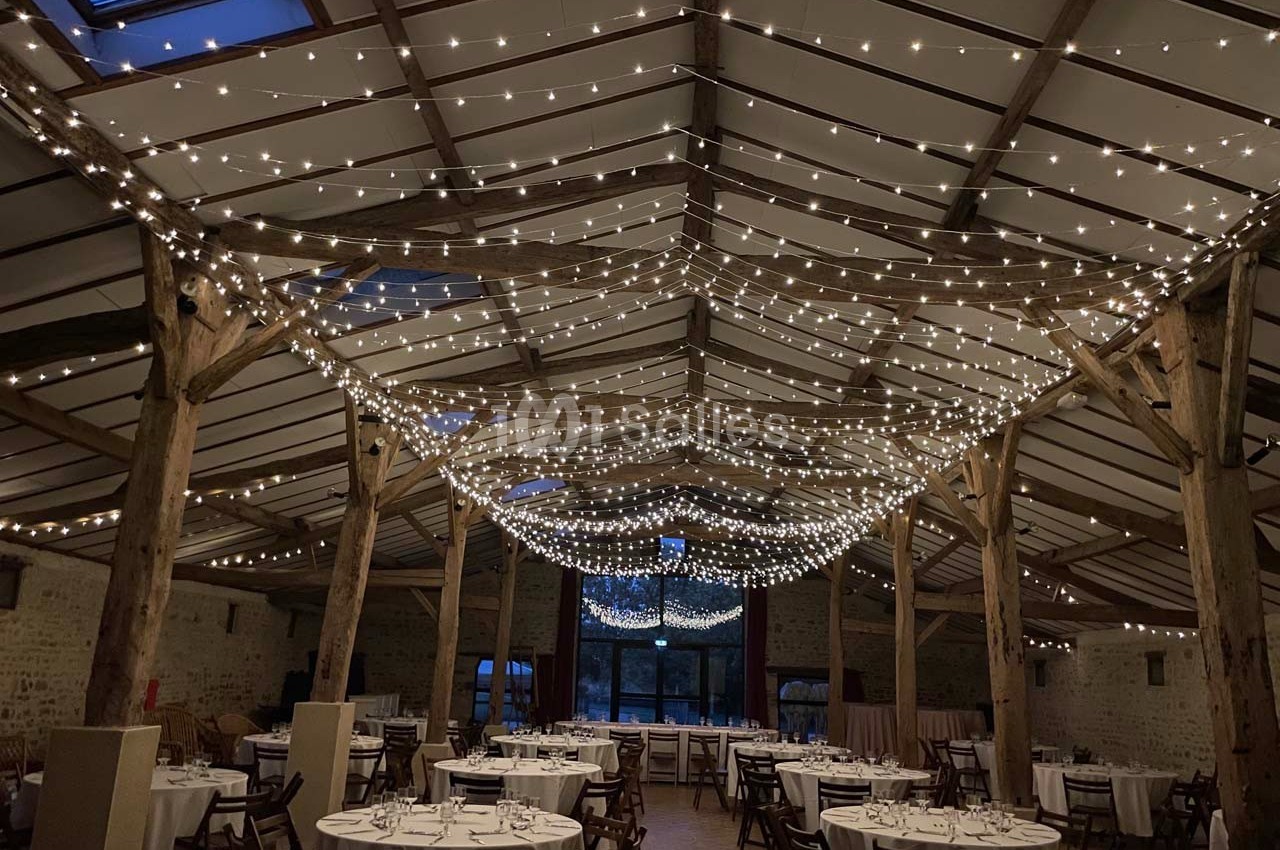 Salle de réception avec tables rondes dressées et guirlandes lumineuses suspendues au plafond en bois.