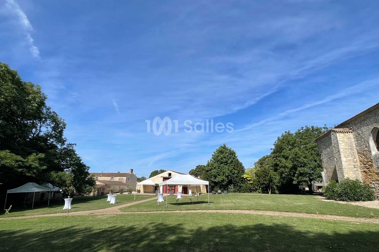 Pelouse avec tentes blanches, tables hautes et bâtiments en pierre sous un ciel bleu dégagé.