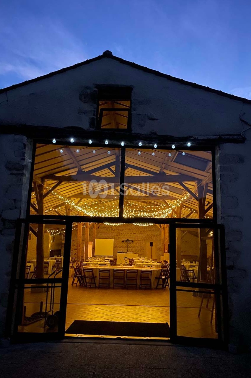 Vue nocturne d'une salle de réception éclairée par des guirlandes lumineuses, visible à travers une grande porte vitrée.