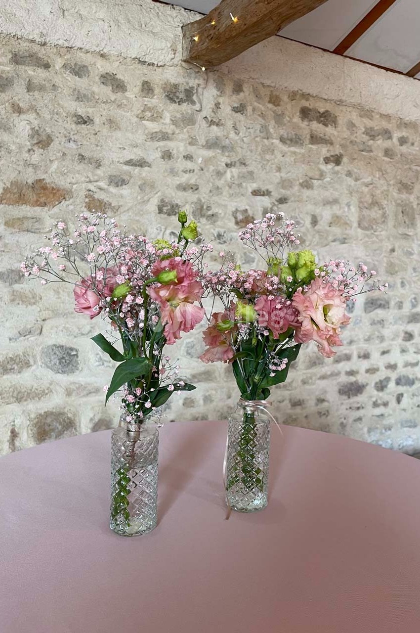 Deux vases en verre contenant des bouquets de fleurs roses et blanches, posés sur une table rose devant un mur en pierre.