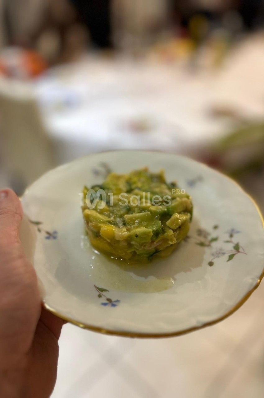Assiette tenue à la main contenant un tartare d'avocat et de mangue, présentée sur une table floue en arrière-plan.
