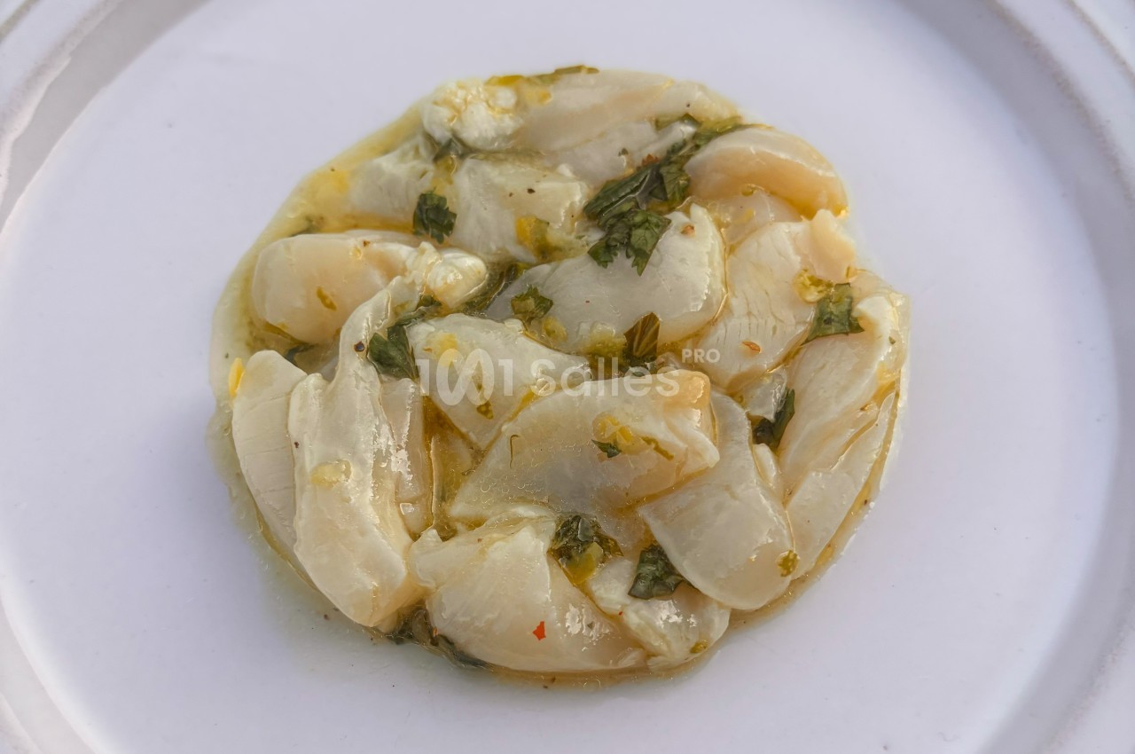 Tranches de noix de Saint-Jacques marinées à l'huile et aux herbes, présentées sur une assiette blanche.