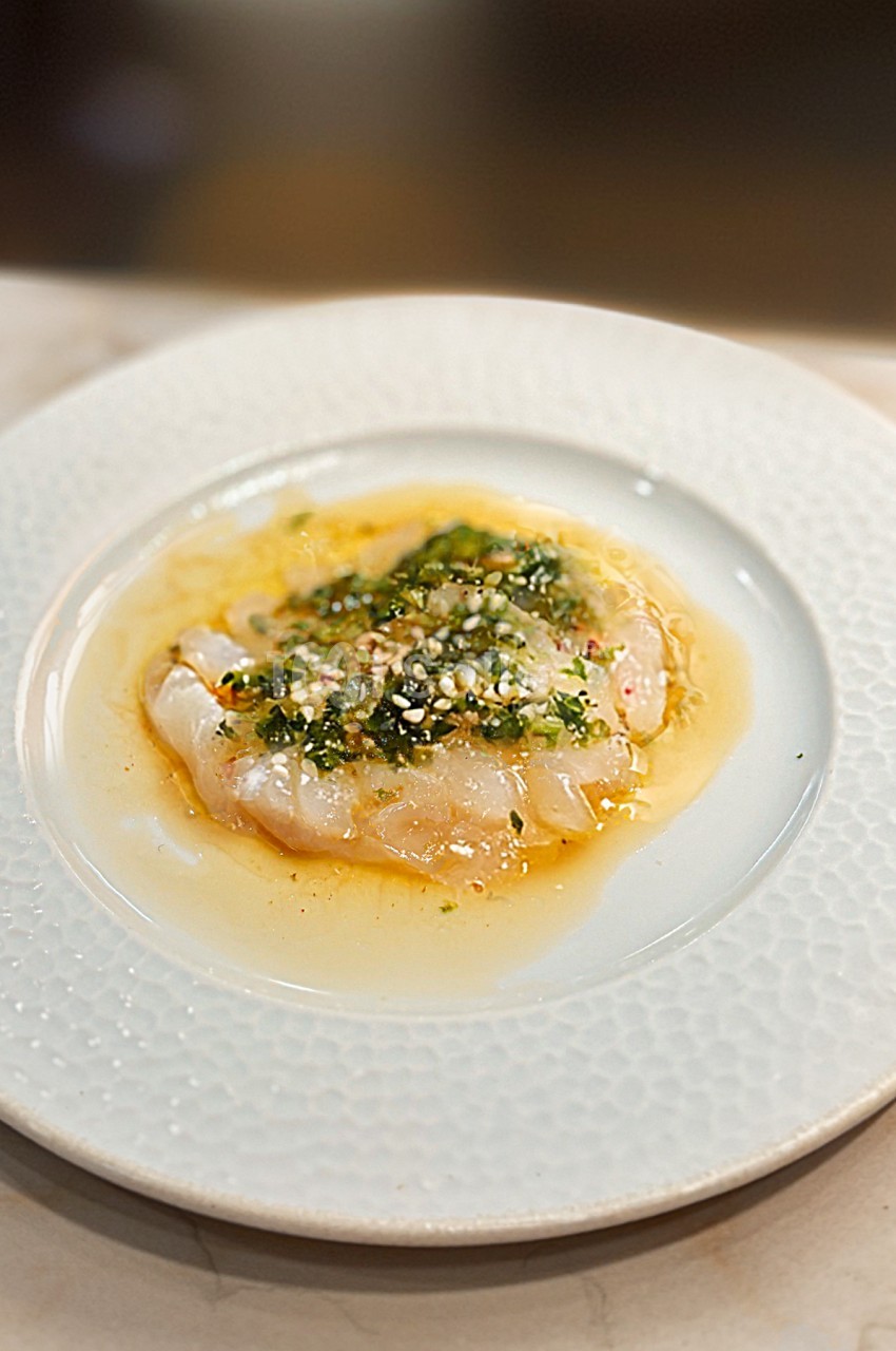 Carpaccio de poisson blanc nappé d'une sauce aux herbes et épices, présenté sur une assiette blanche texturée.