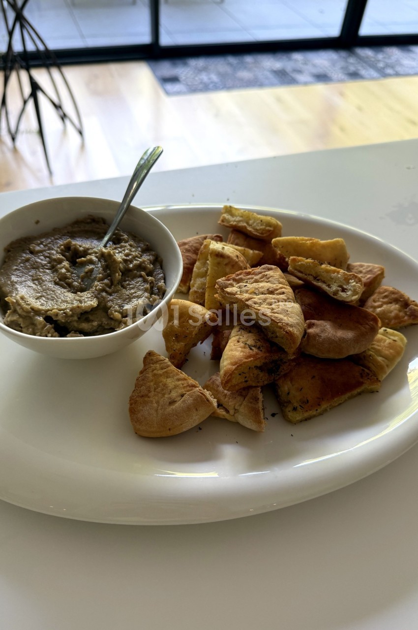 Assiette de pita grillée accompagnée d'un bol de houmous, posée sur une table blanche près d'une baie vitrée.