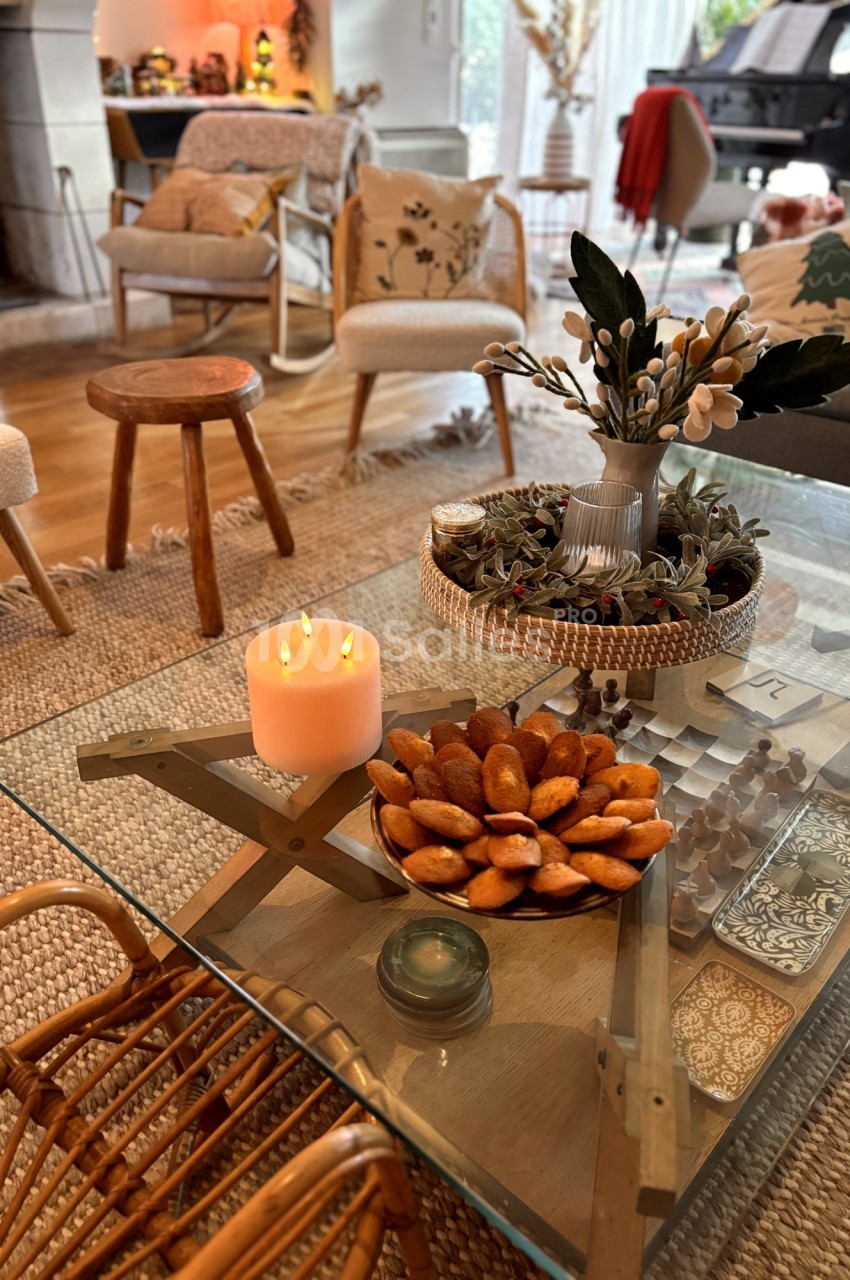 Table basse en verre décorée d'une bougie allumée, d'un plat de madeleines et d'un vase avec des branches, dans un salon…