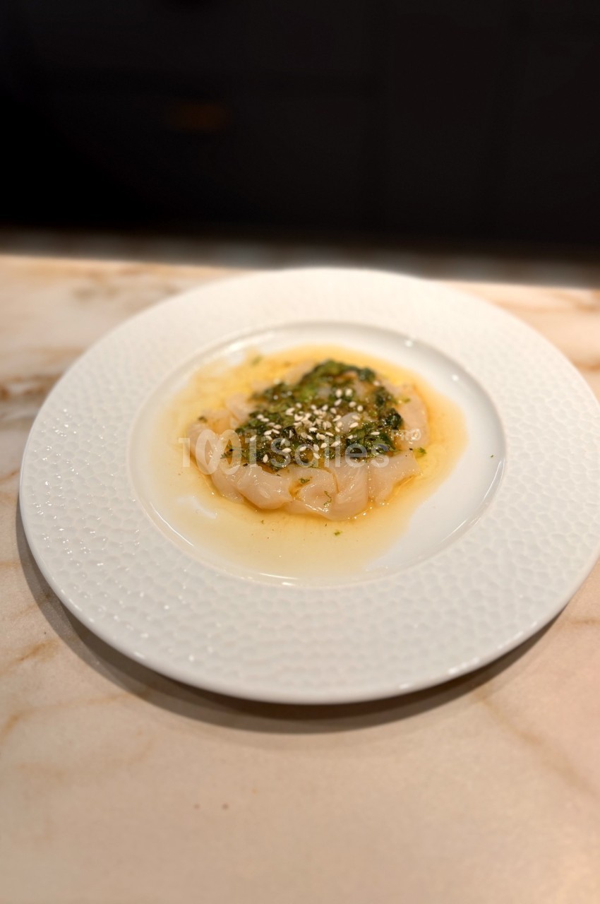 Assiette de carpaccio de poisson blanc avec sauce verte et graines de sésame, servie sur une table en marbre.