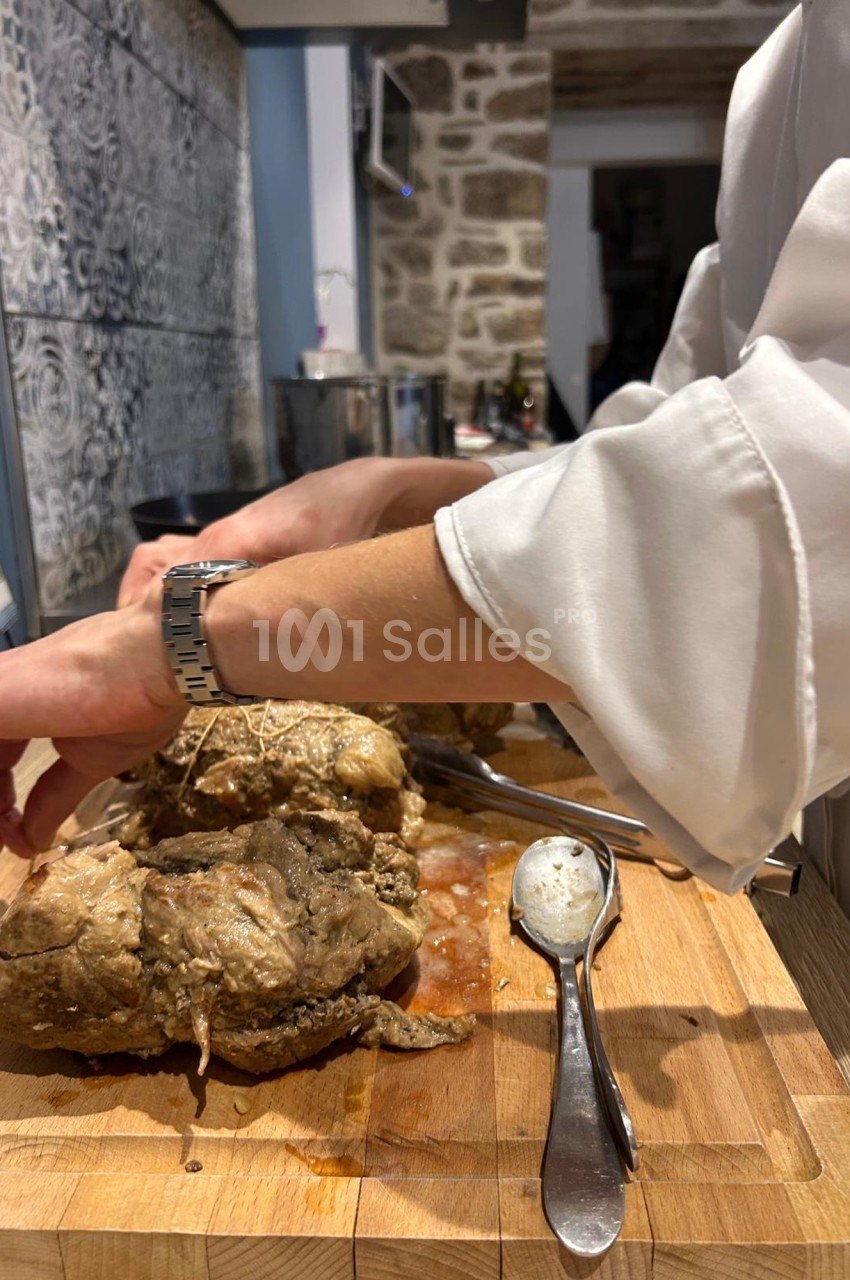 Un cuisinier attache une pièce de viande cuite sur une planche en bois, entourée d'ustensiles de cuisine.