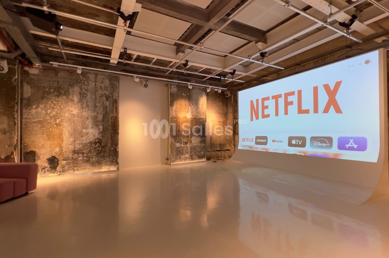 Salle moderne avec un grand écran affichant le logo Netflix et des icônes de plateformes de streaming, murs bruts en arrière…