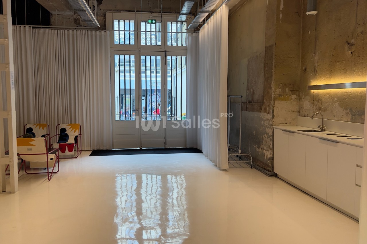 Espace intérieur lumineux avec sol blanc, murs bruts, kitchenette moderne et grandes portes vitrées donnant sur l'extérieur.
