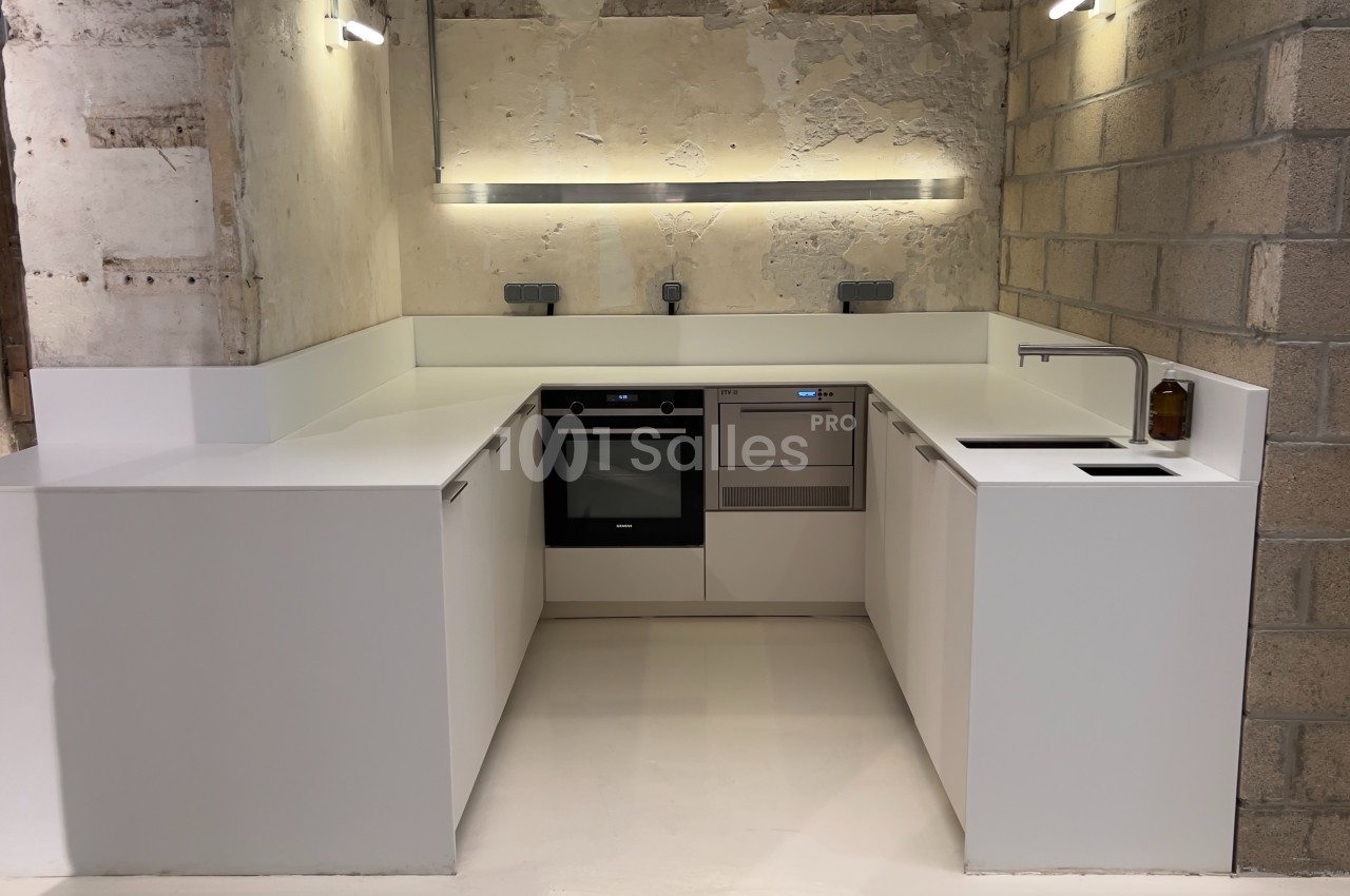 Cuisine moderne en U avec meubles blancs, plan de travail épuré, mur en béton brut et éclairage intégré.