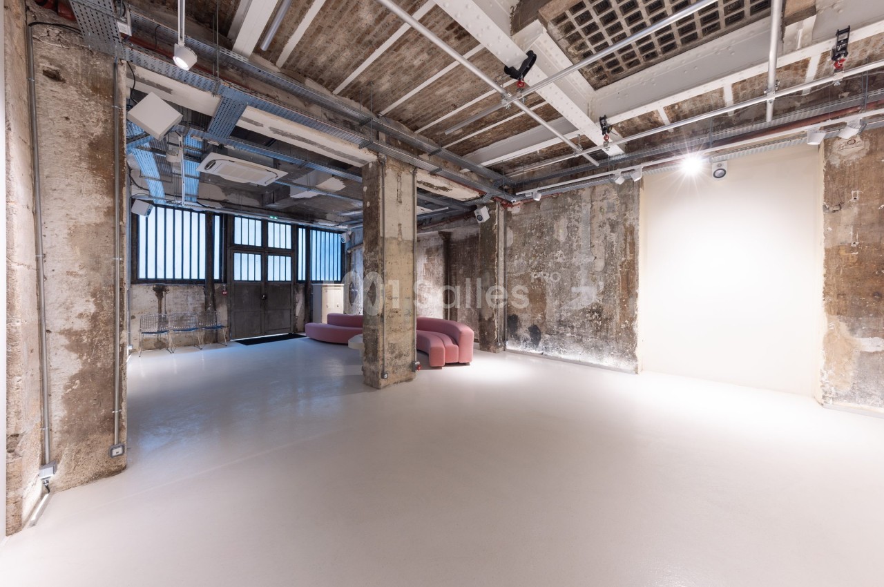 Espace intérieur industriel avec murs en béton brut, sol clair, canapé rose et grandes fenêtres à barreaux.