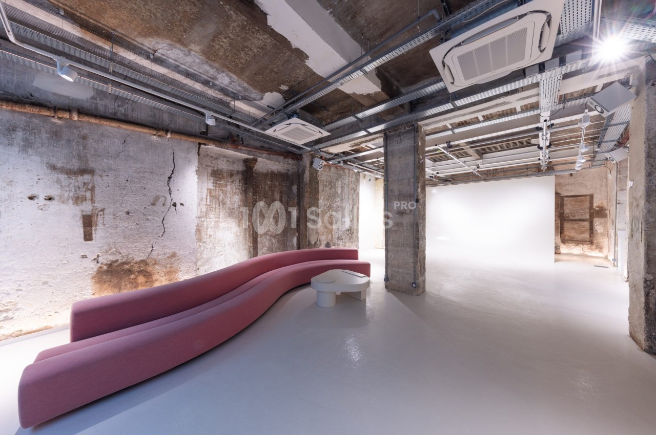 Espace intérieur industriel avec murs bruts, plafond apparent et canapé rose incurvé sur un sol blanc.