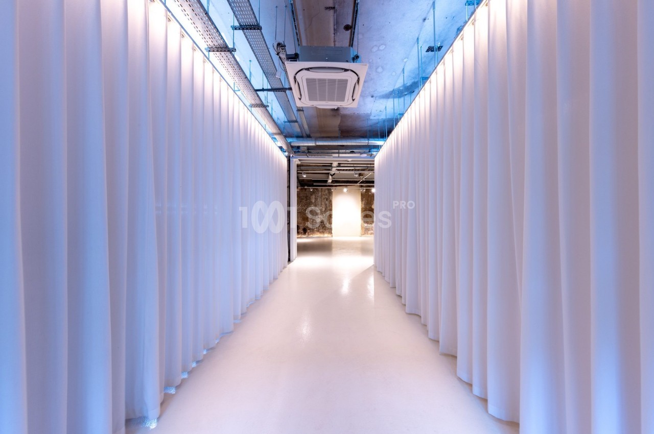 Couloir lumineux avec sol blanc, rideaux blancs de chaque côté et plafond industriel exposant des conduits.