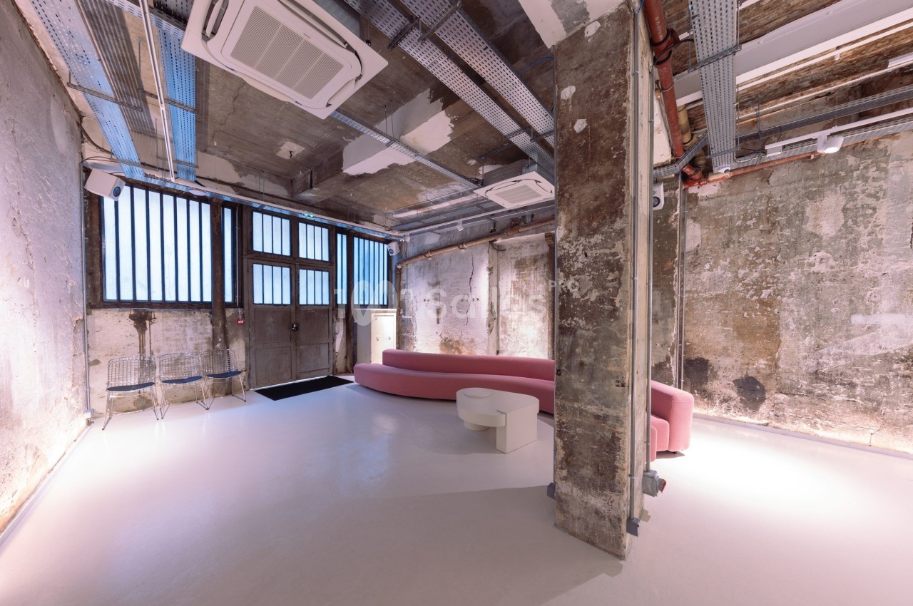 Espace intérieur industriel rénové avec murs bruts, canapé rose, chaises transparentes et lumière naturelle.