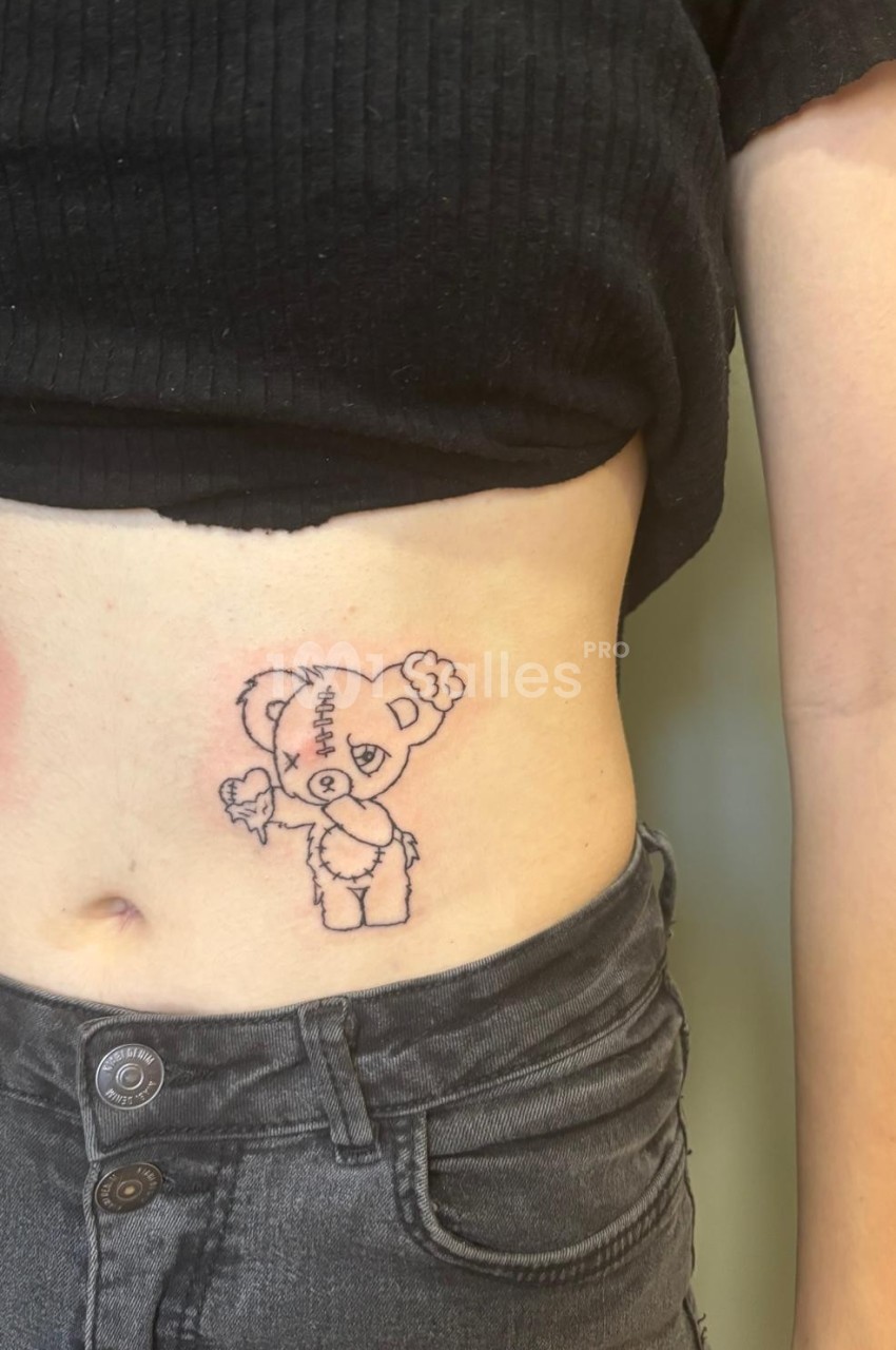 Tatouage d'un ours en peluche avec des détails de couture, situé sur le côté de l'abdomen d'une personne.